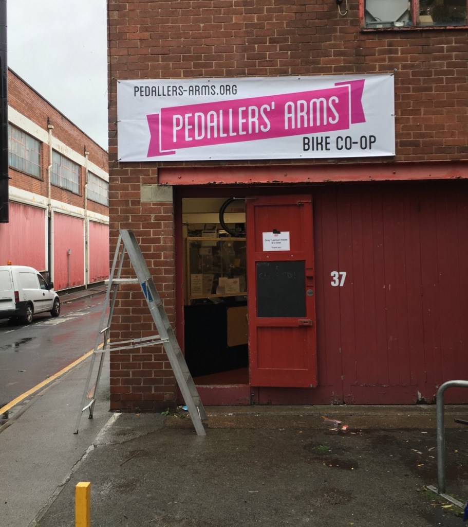 Photo of the entrance to the Pedallers' Arms workshop. There is a banner with the logo mounted on a brick building above red doors. Banner has pink logo on white background. There is a step ladder on the ground.
